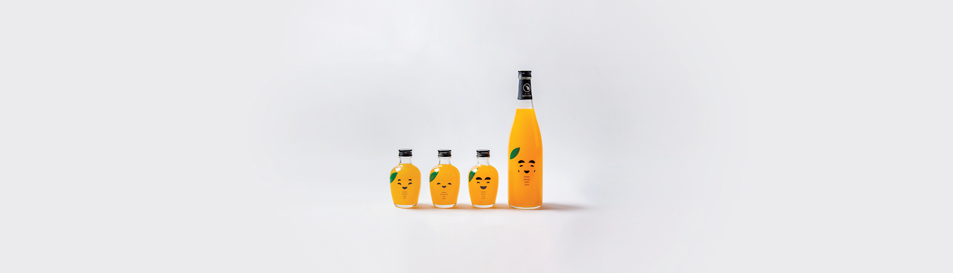 Quatro garrafas de vidro transparente com rostos desenhados nela, com líquido laranja dentro, fazendo alusão à fruta laranja. O fundo da imagem é branco.