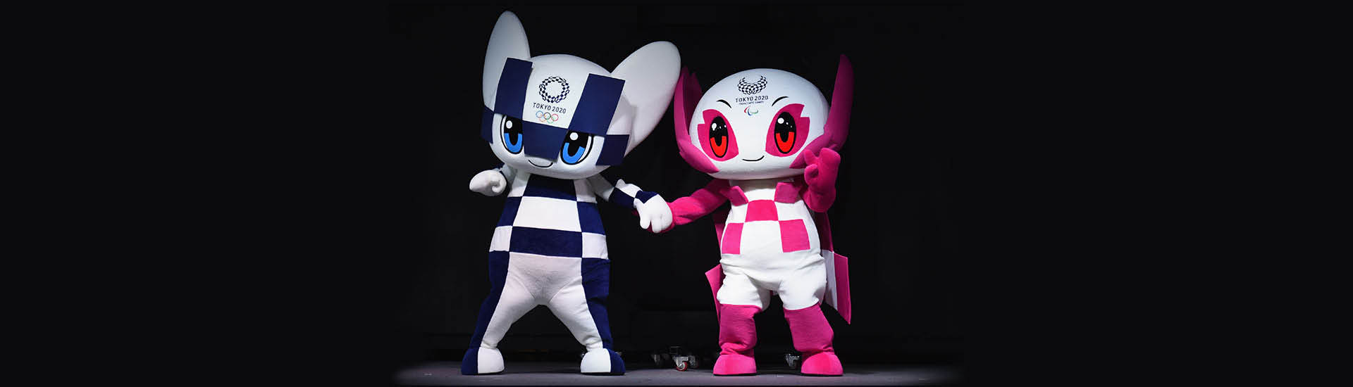 Mascotes dos Jogos Olímpicos e Paralímpicos de Tokyo 2020. À esquerda, Miraitowa: personagem com roupa branca com quadriculados azuis, olhos grandes e azuis, com orelhas para cima. À direita, Someity: personagem com roupa branca e quadriculados rosas, olhos vermelhos e capa rosa. O fundo da imagem é preto.