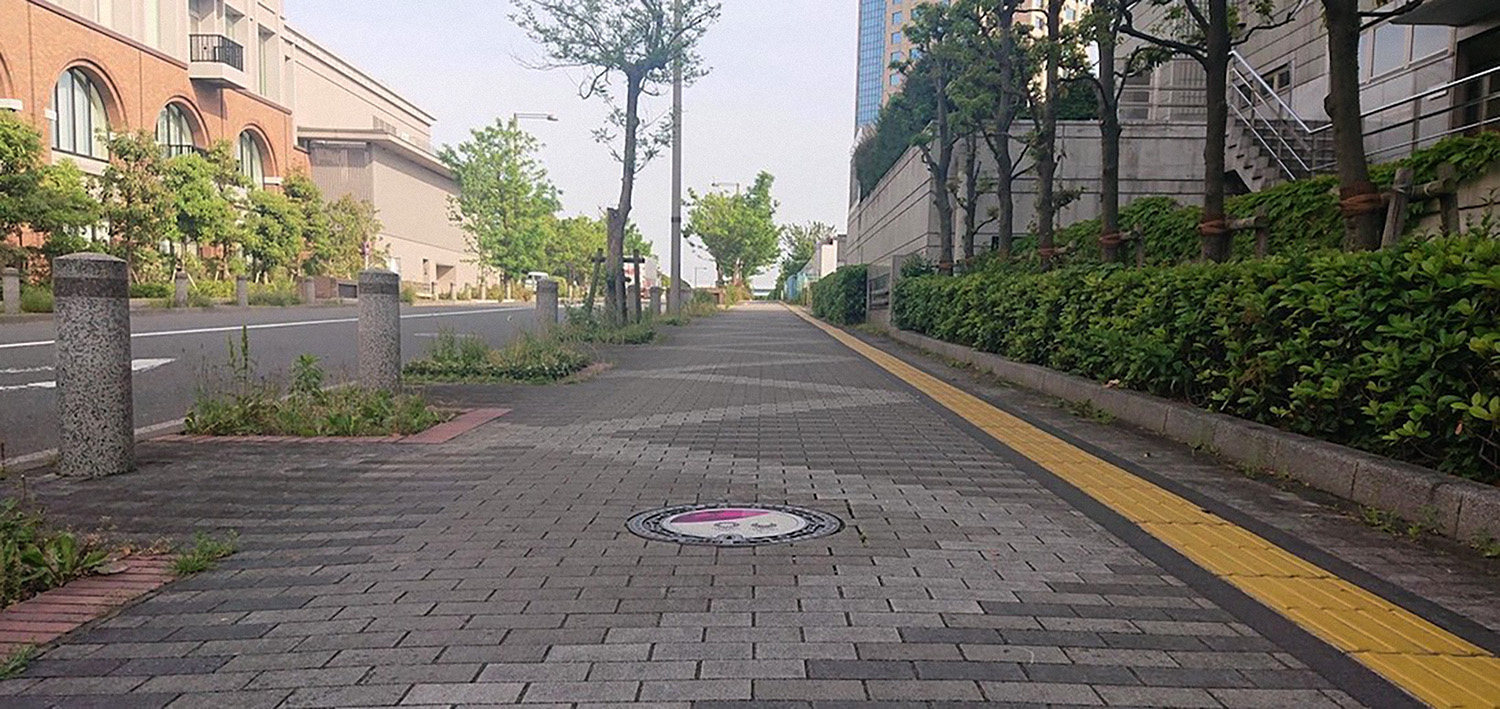 Foto de rua no Japão com uma tampa de bueiro no meio da calçada estampada com o tema dos Jogos Olímpicos de Tokyo 2020 no chão.