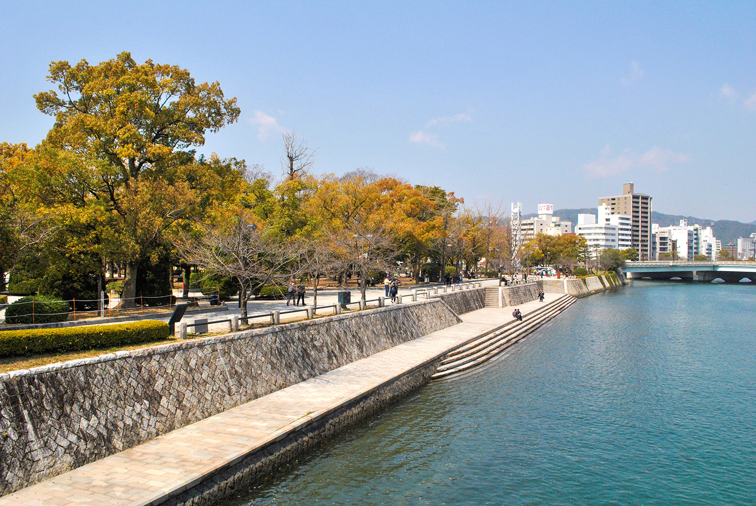 Parque do Memorial da Paz. Foto da margem de um rio com árvores com folhagens verde e amarela e céu azul. 