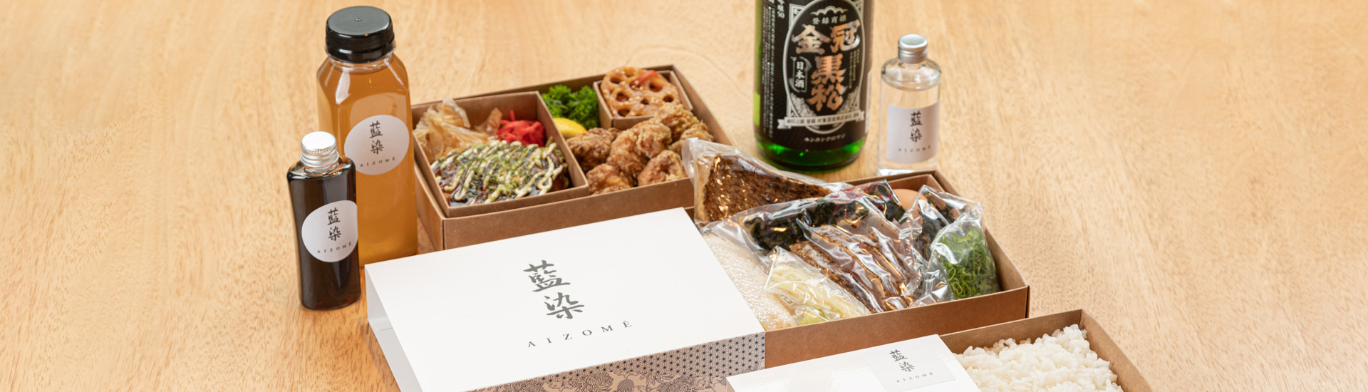 Kit gastronômico da Experiência JHSP: Cultura e Gastronomia de Setouchi Oeste. Garrafas de diferentes tamanhos e caixas de comidas sobre mesa de madeira clara. 