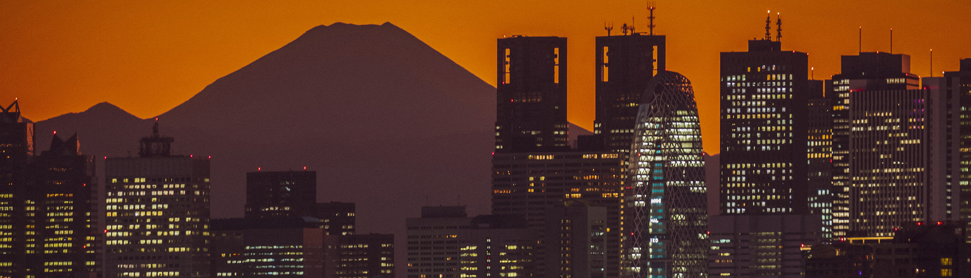 Cena urbana com diversos prédios ao entardecer, com o céu alaranjado e a silhueta do  Monte Fuji ao fundo.