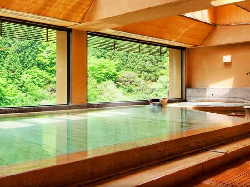 Onsen - termas de água aquecida - dentro de uma sala de madeira, com janelas de vidro que mostram uma bela paisagem de natureza verde.
