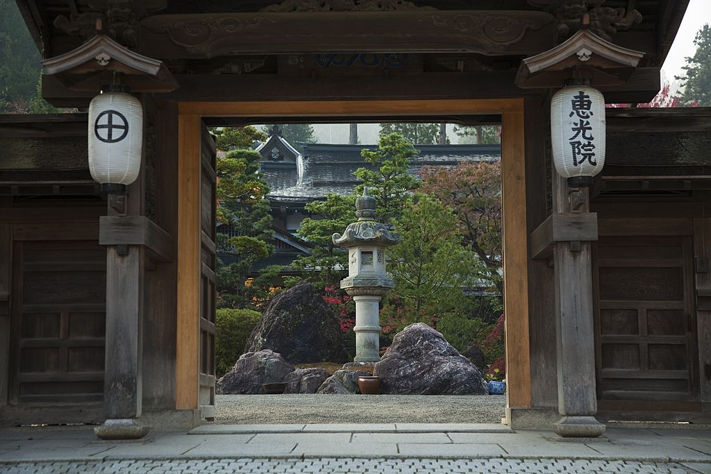 Entrada de hospedagem estilo ryokan, com muro e portão de madeira, por onde é possível avistar um jardim japonês.