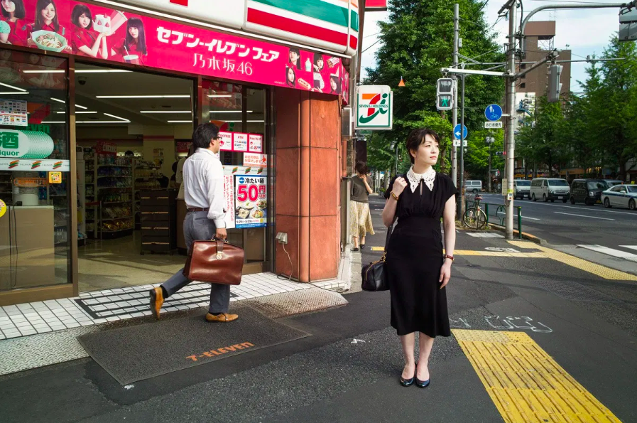 A autora japonesa Sayaka Murata está em pé, com um vestido branco, em frente a uma loja de conveniência, uma konbini.