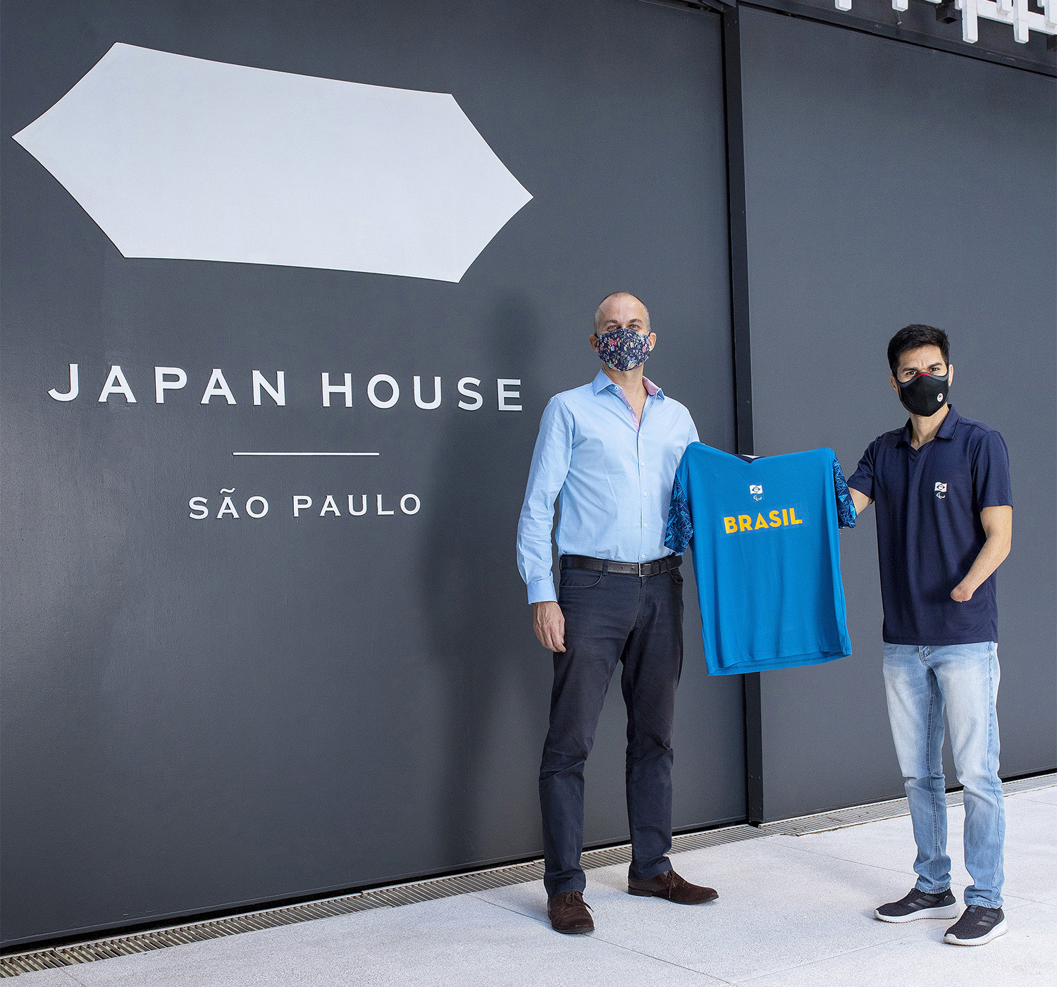 Eric Klug, presidente da Japan House São Paulo, com o ex-atleta do atletismo Yohansson Nascimento, vice-presidente do Comitê Paralímpico Brasileiro, segurando uma camisa azul em frente à Japan House São Paulo