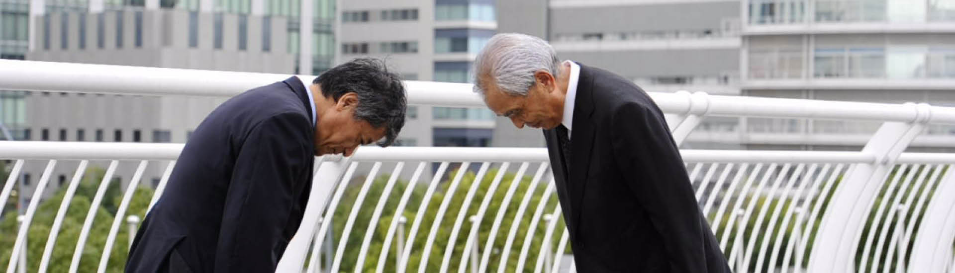 Dois homens de terno preto se cumprimentam com gesto japonês de se curvar, abaixando a cabeça, em demonstração de respeito.