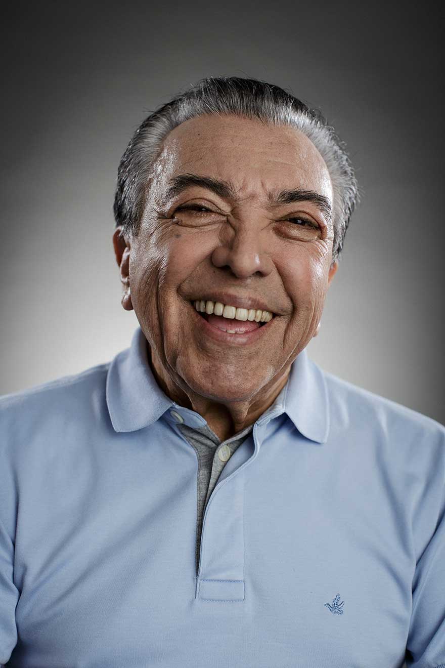 Retrato de Mauricio de Sousa, vestindo uma camisa polo azul clara, sorrindo para a câmera. O fundo da imagem é cinza.