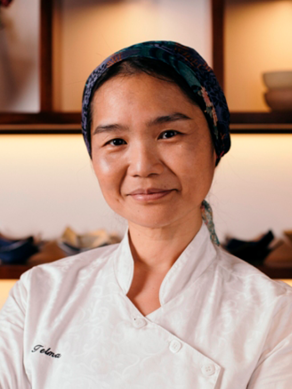 Telma Shiraishi. Mulher com traços asiáticos, pele clara, cabelo preso com um lenço, vestindo  um uniforme branco de chef de cozinha.