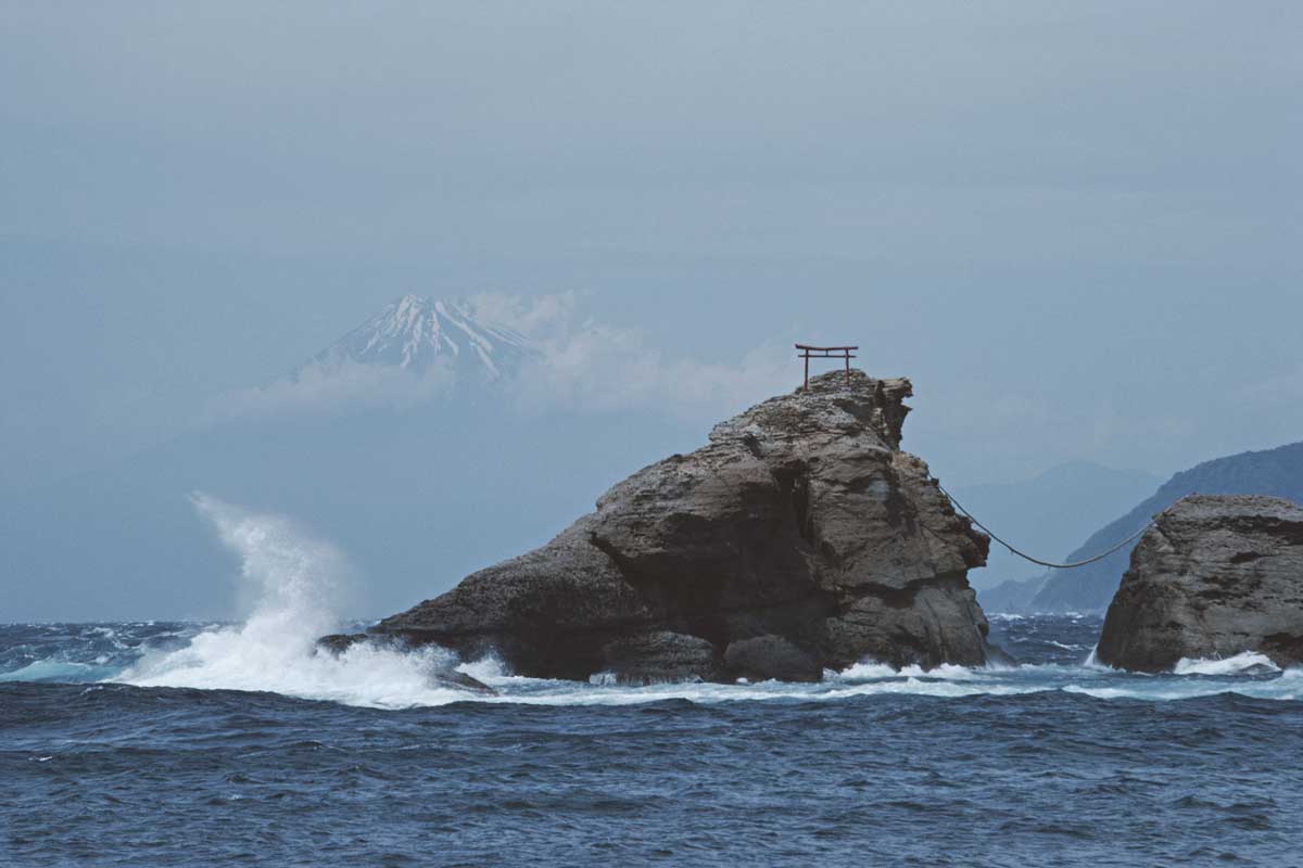 Onda batendo em rocha no meio do mar azul escuro. Ao fundo é possível enxergar a silhueta do Monte Fuji, no Japão, em meio às nuvens,