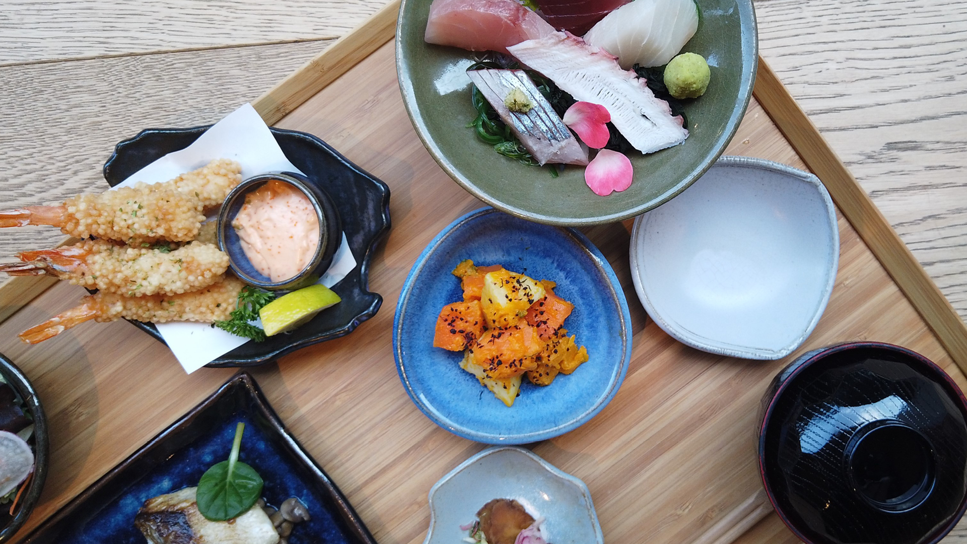 Bandeja de madeira com diversos recipientes coloridos de cerâmica com pratos japoneses, como camarão empanado, peixe, conservas, etc.