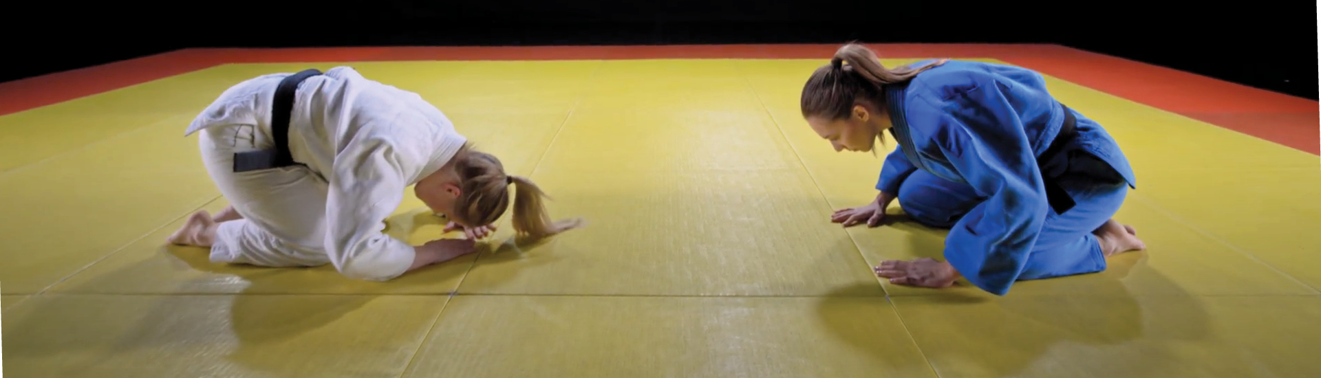 Duas atletas de artes marciais se curvam, ajoelhadas no chão, uma de frente para a outra, em sinal de respeito. Vestem kimonos tradicionais do esporte, nas cores branca - à esquerda - e azul - à direita. O chá é amarelo com bordas alaranjadas.