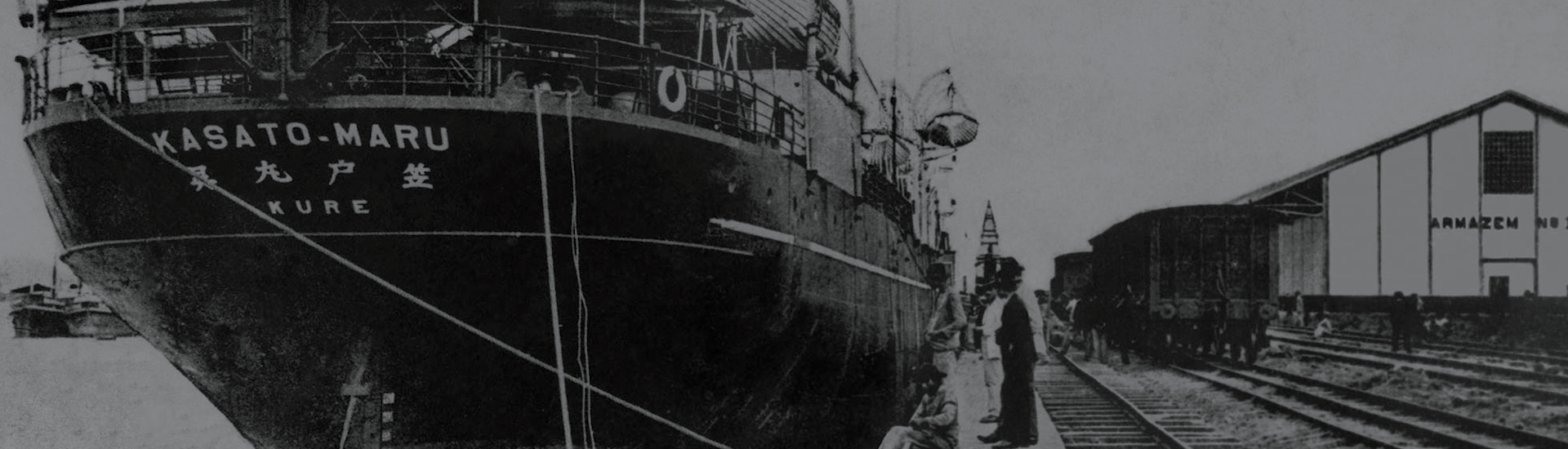 Foto em preto e branco, mostrando a proa do navio Kasato Maru, ao lado de um trilho de trem e um grande galpão escrito "Armazem".