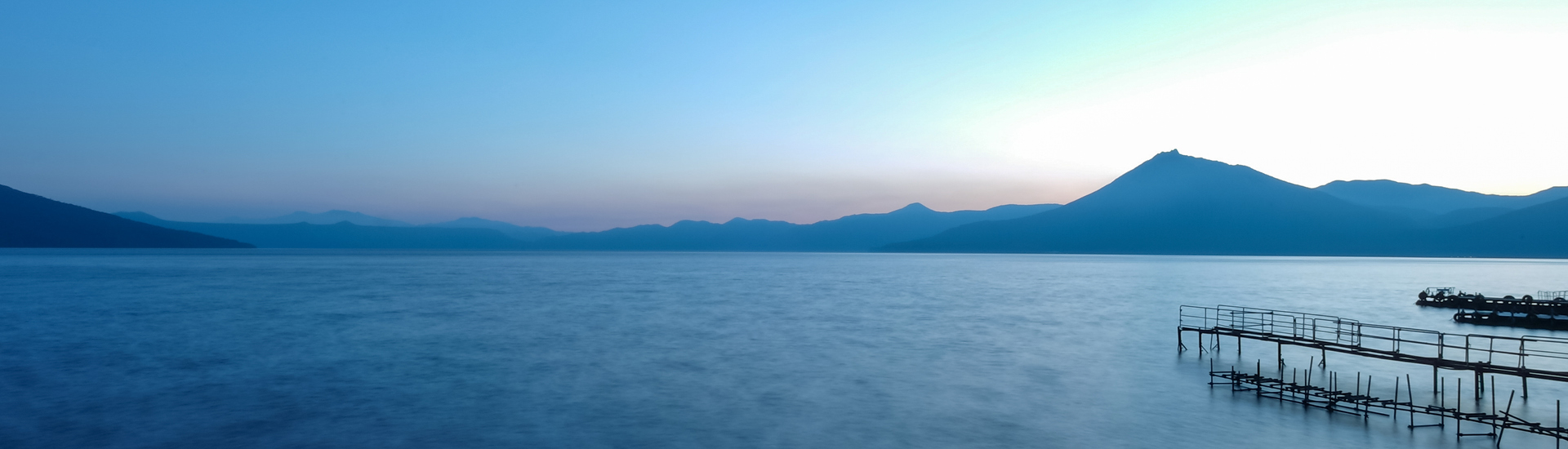Mar azul com montanhas no horizonte e um deque à direita da foto.