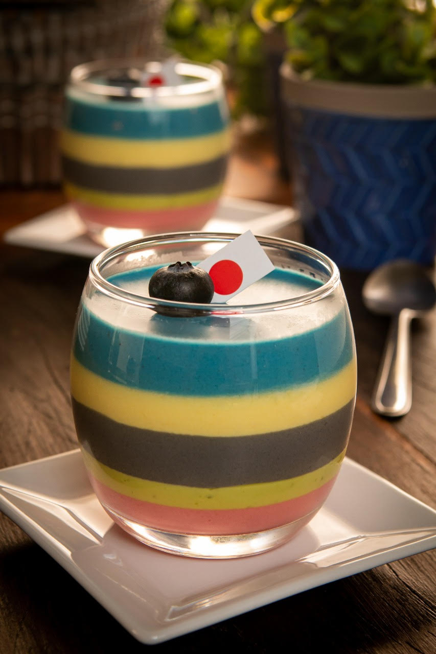Pote de vidro com mousse em camadas coloridas, enfeitado no topo com uma frutinha mirtilo e uma mini bandeira do Japão.