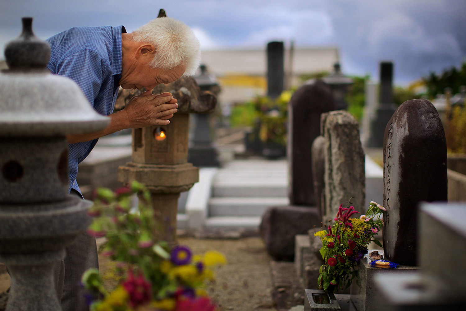 Homem japonês, de cabelos curtos brancos, curvado com as mãos em oração em um cemitério. Ao fundo, céu azul.