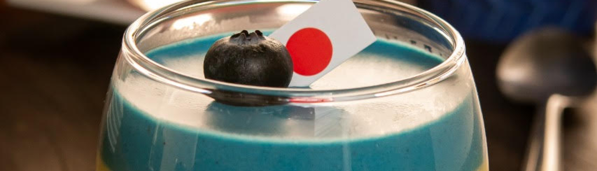 Pote de vidro com mousse azul, enfeitado no topo com uma frutinha mirtilo e uma mini bandeira do Japão.
