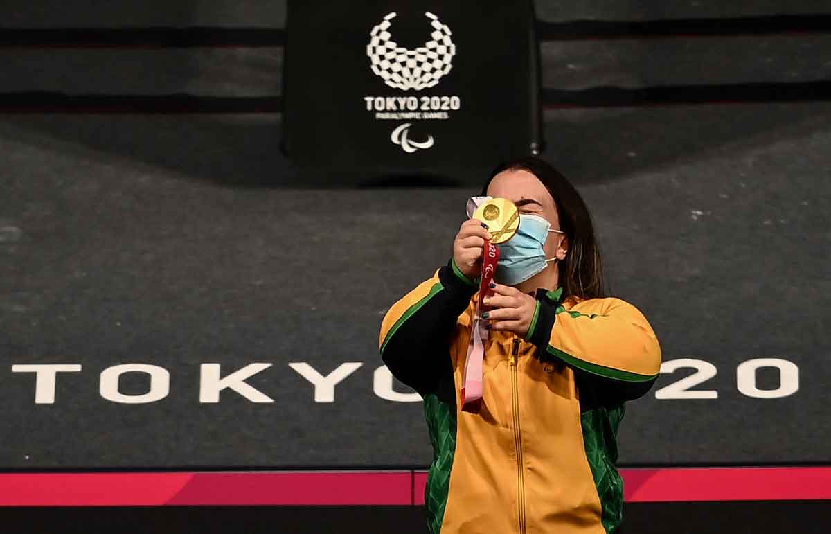 Atleta halterofilista Mariana D'Andrea, com jaqueta amarela e verde, levanta a medalha de ouro em frente de seu rosto. Atrás dela é possível ver um carpete preto com o escrito TOKYO 2020 em letras brancas e símbolo dos Jogos Paralímpicos ao fundo.