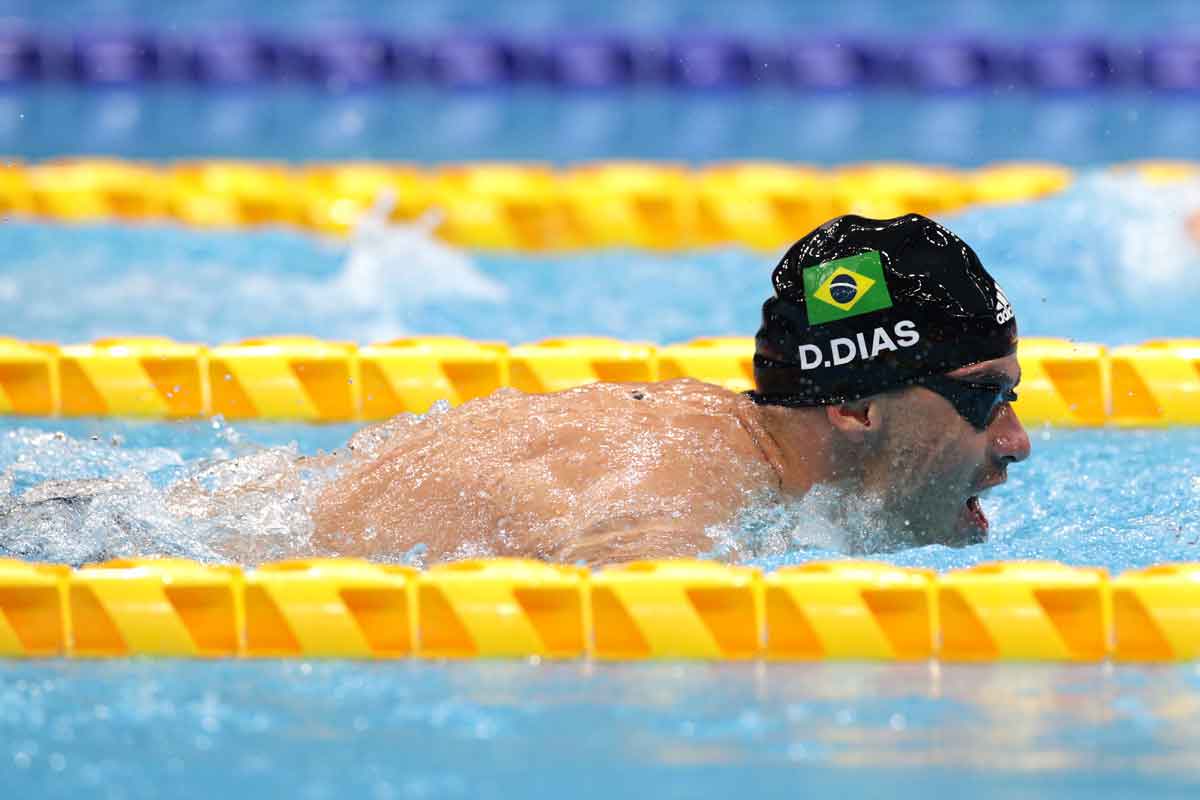 Atleta Daniel Dias, nadando em piscina azul com divisórias amarelas, com touca preta com bandeira do Brasil e o escrito "D. Dias" em letras brancas.