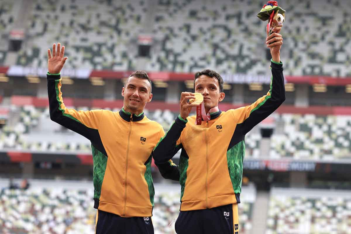 Dois atletas de atletismo, Yeltsin Jacques e o atleta-guia, com jaqueta amarela e verde, de braços levantados acenando para arquibancada com medalha de ouro no peito. Ao fundo, arquibancada de estádio desfocada.
