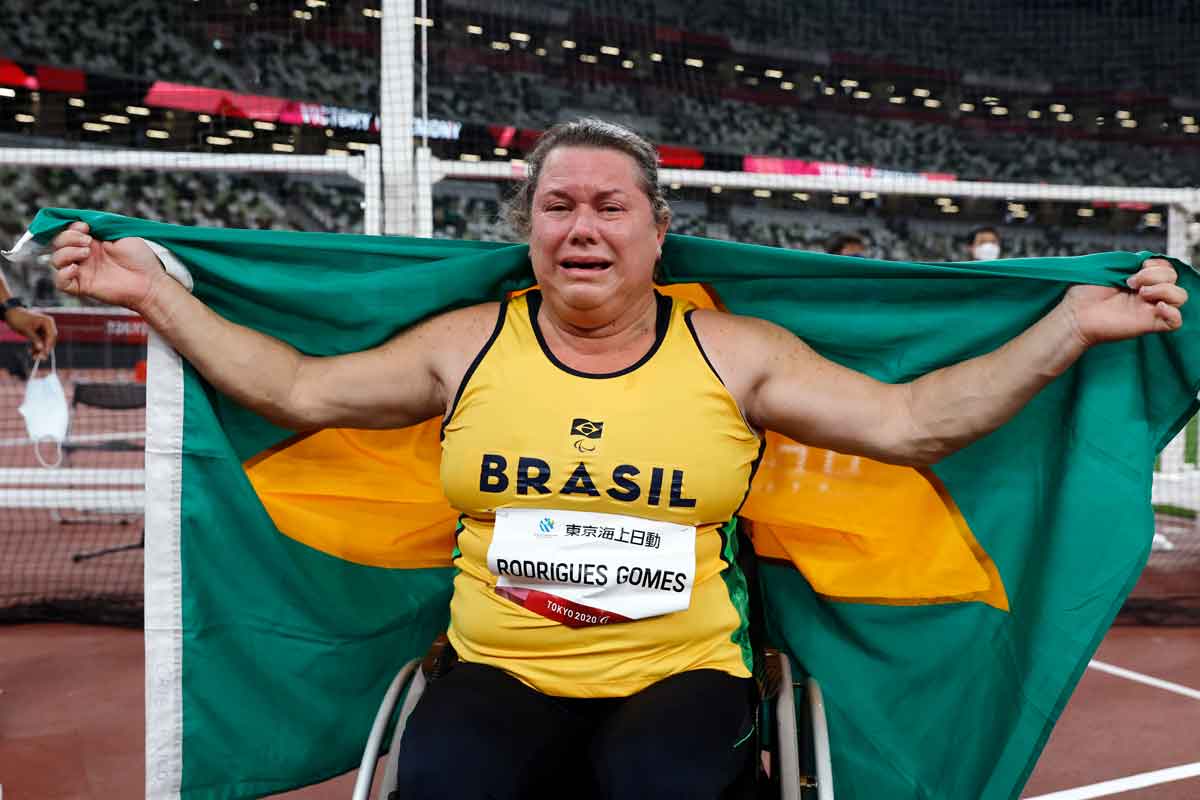 Beth Gomes, atleta de lançamento de disco, com regata amarela sentada em cadeira de rodas, segura bandeira do Brasil emocionada. 
