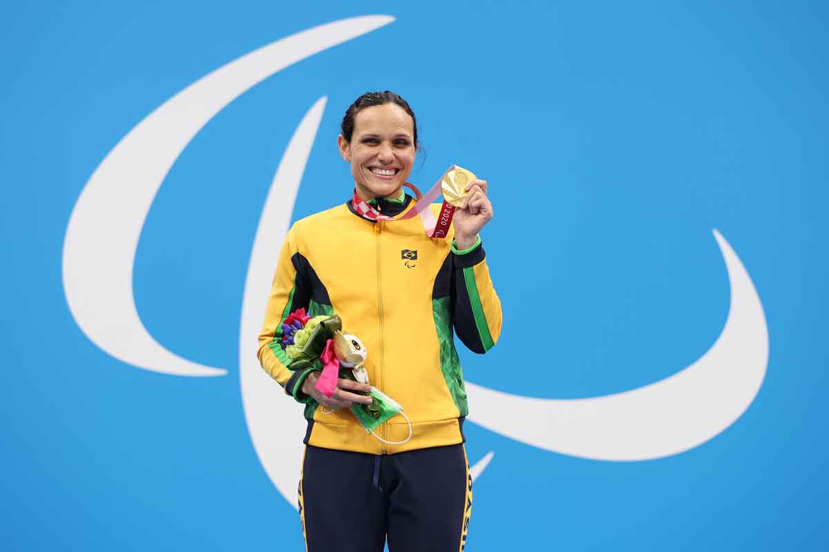 Atleta Carol Santiago, da natação, com jaqueta amarela e verde, segura medalha de ouro em uma mão e mascote paralímpica em outra. Atrás dela, símbolo dos Jogos Paralímpicos em branco com fundo azul claro.