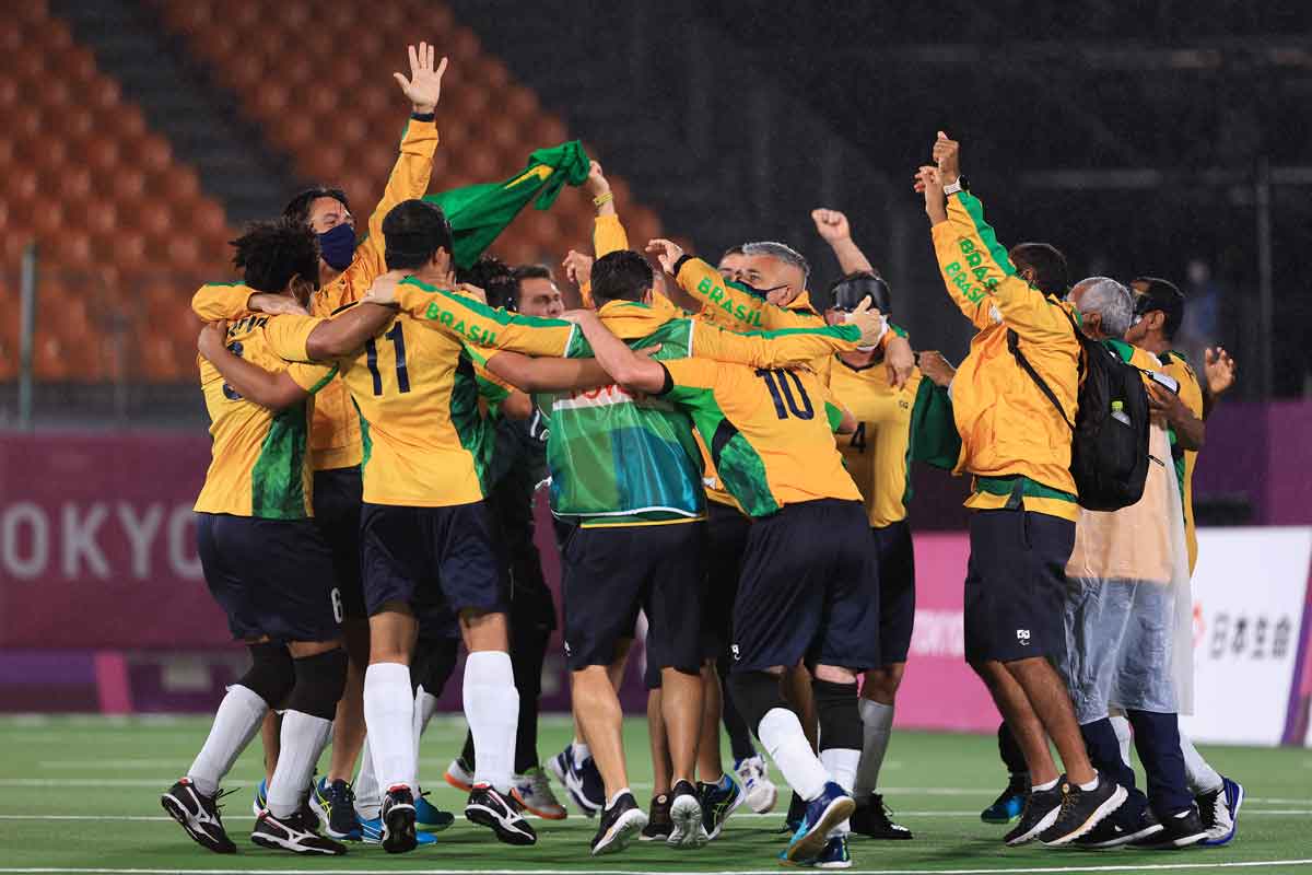 Atletas do Futebol de 5, com jaqueta amarela e bermuda preta, se abraçam comemorando vitória em campo de futebol. Ao fundo, arquibancada em desfoque.