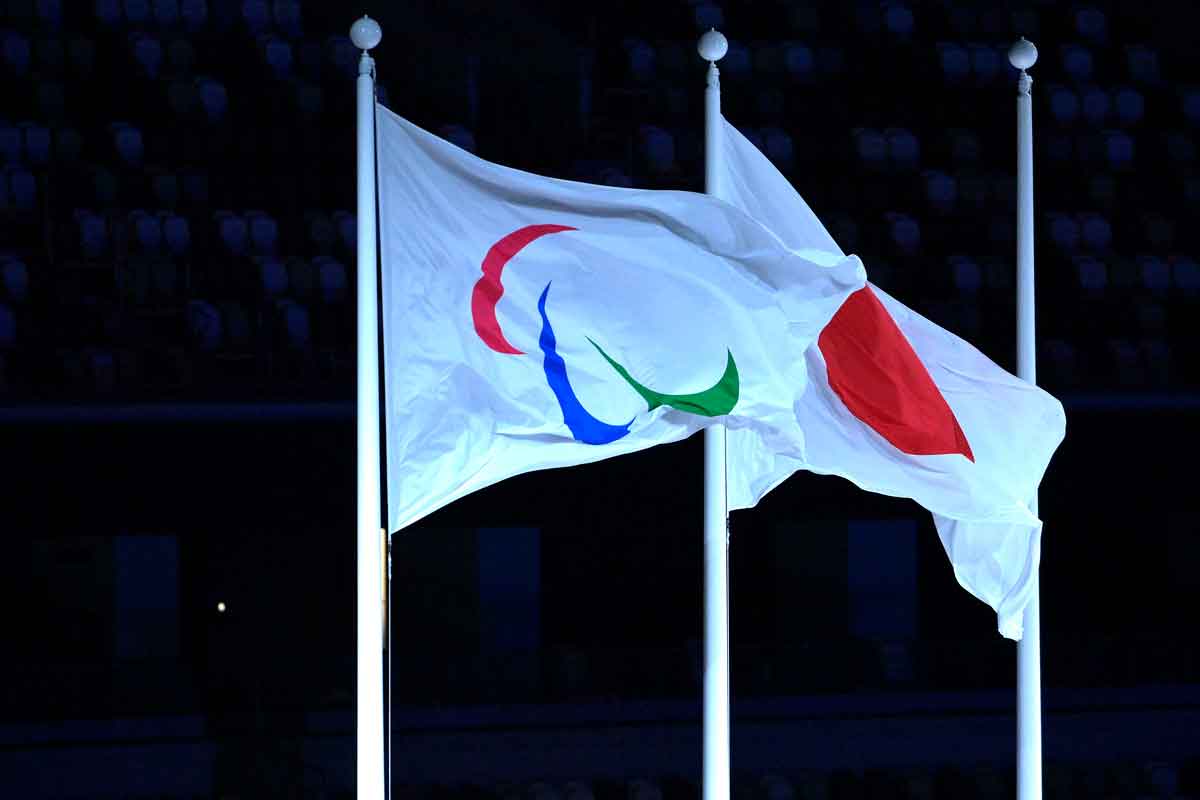 Bandeira com símbolo dos Jogos Paralímpicos e bandeira do Japão, hasteadas lado a lado. O fundo da imagem é preto.