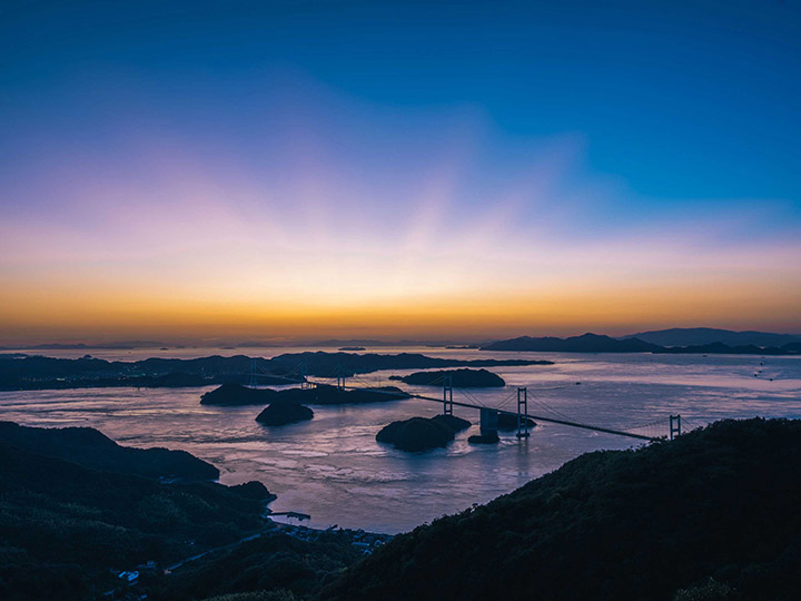 Setouchi. Vista de diversas ilhas com céu azul e sol nascendo no horizonte.
