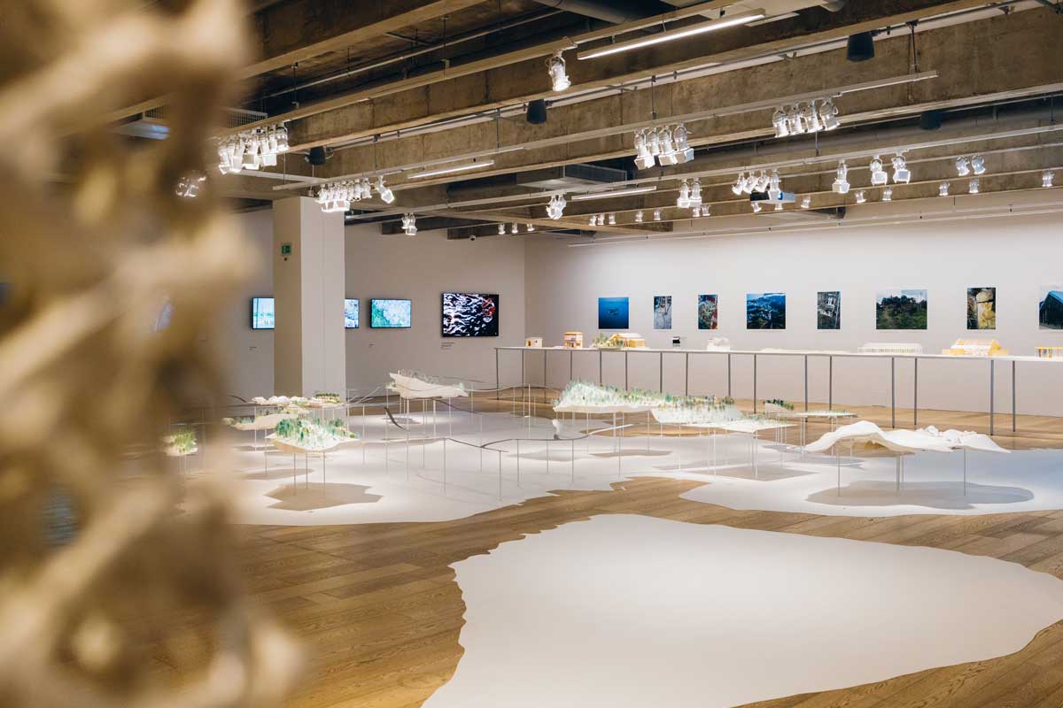 Espaço expositivo de piso de madeira com adesivos brancos em formato orgânico e paredes brancas. Exposição “Simbiose: a ilha que resiste”, no 2º andar da JHSP.