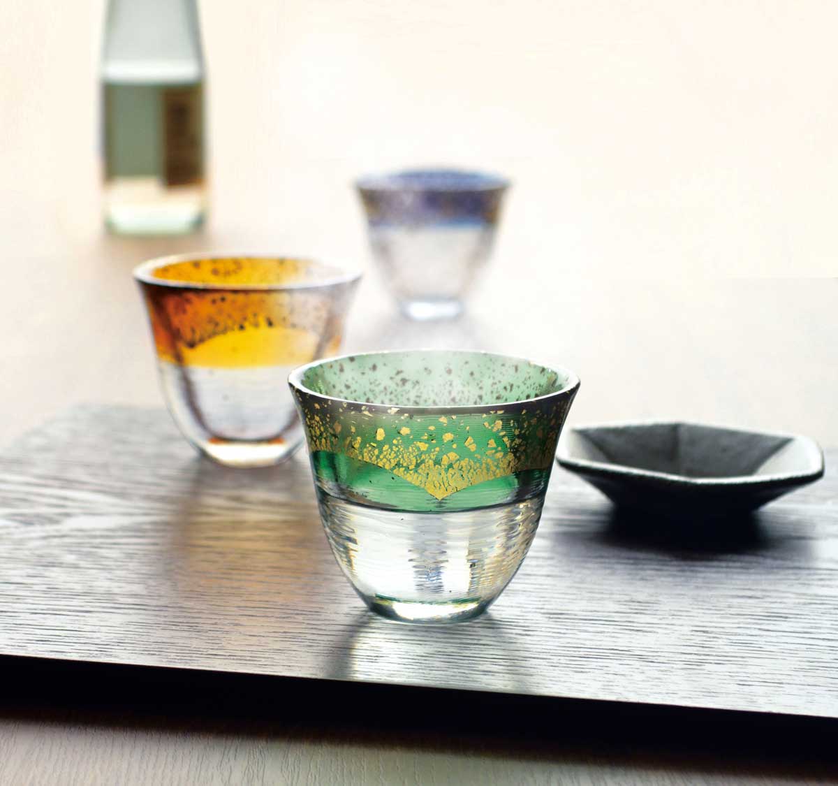 Foto de três copos de vidro transparente com borda colorida e pequeno recipiente preto sobre mesa de madeira.