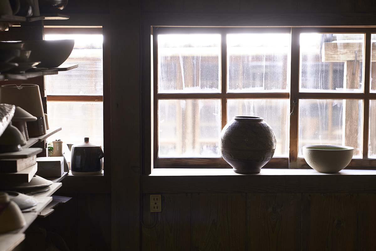 Peças de cerâmica expostas em uma estante e bancada de madeira em um cômodo com uma grande janela.
