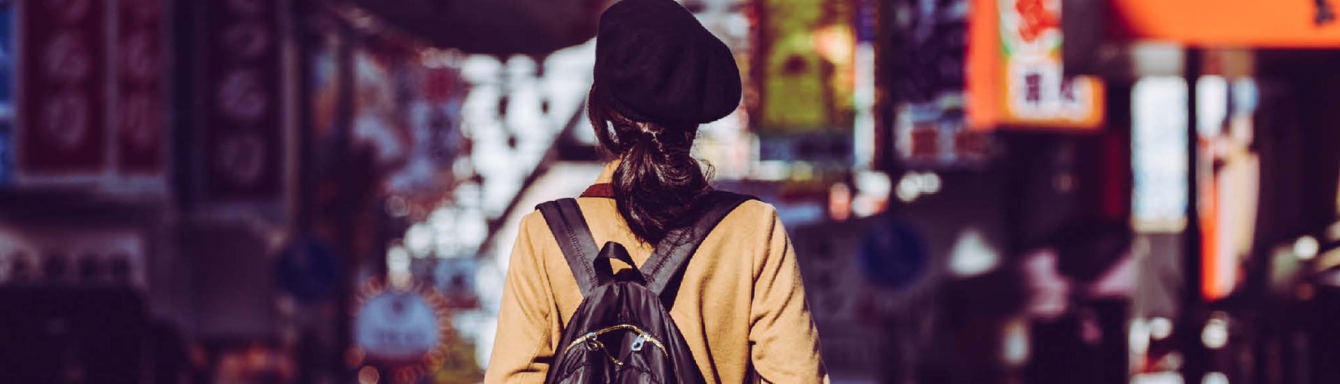 Foto de uma garota de costas vestindo um casaco mostarda, boina preta e mochila em uma paisagem urbana.
