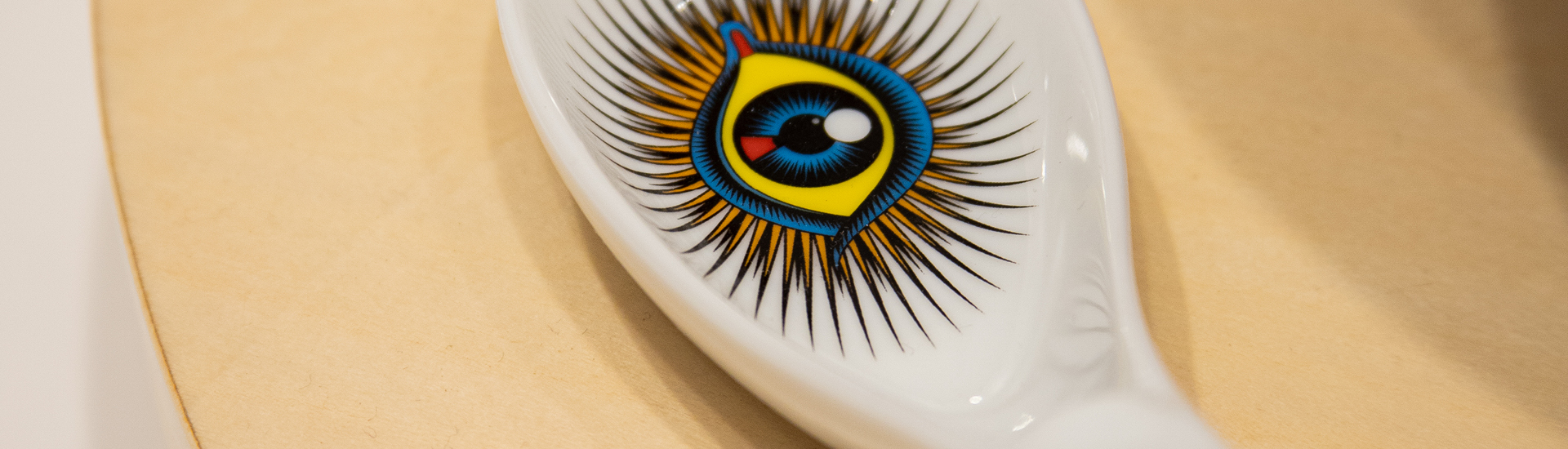 detalhe de colher de cerâmica branca com desenho de um olho colorido, sobre mesa branca