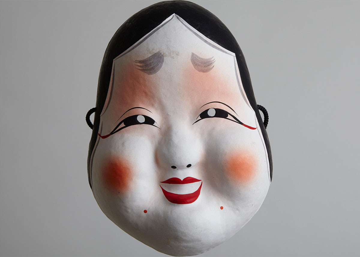  Imagem de uma máscara branca e sorridente de  Otafuku, ou Okame, às vezes chamada de Deusa da Hilaridade.