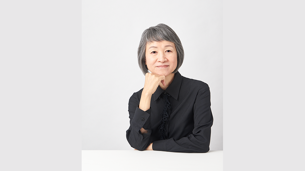  Fotografia de Reiko Sudo. Reiko é uma mulher grisalha, de cabelos curtos e traços asiáticos. Ela olha para frente com um leve sorriso, e veste um terno preto.