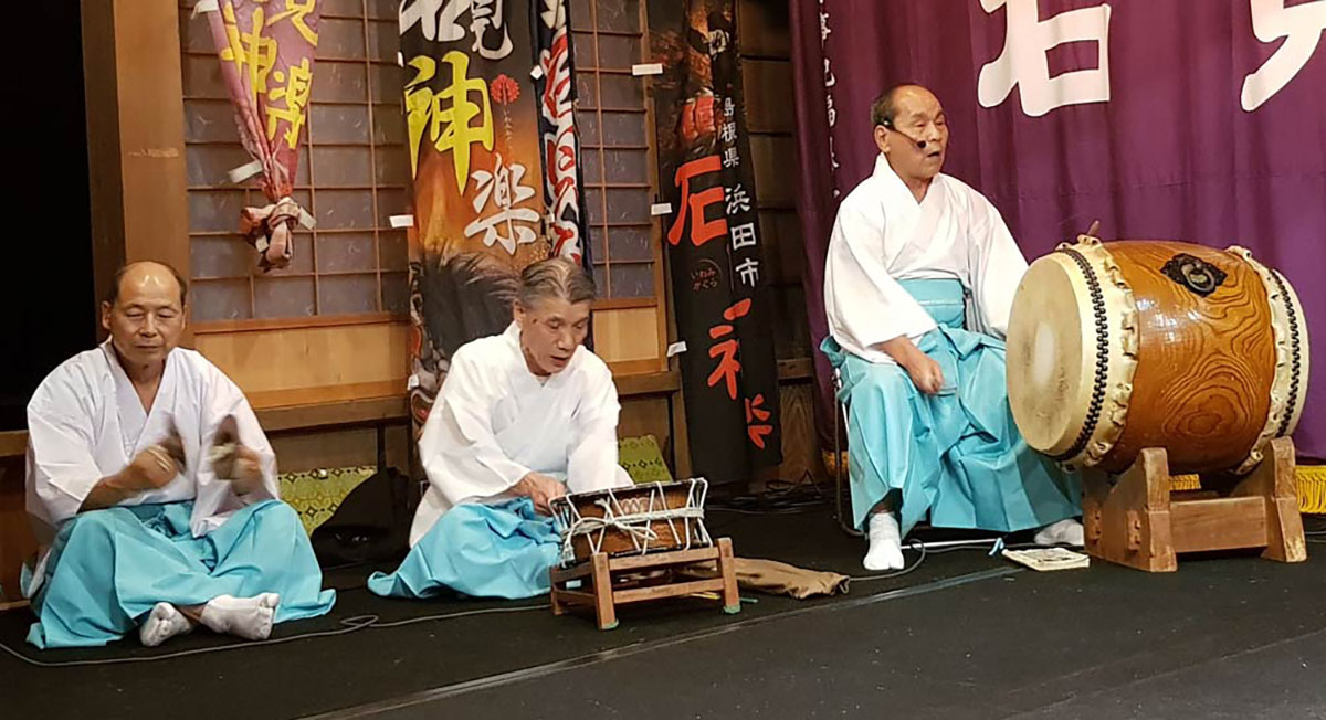 Imagem de homens, com traços asiáticos, ornamentados em uma apresentação tocando um instrumento típico