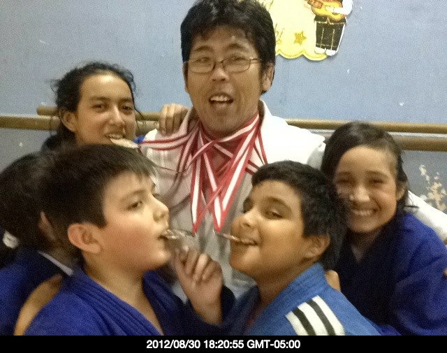 Fotografia do judoca Futoshi Urata com diversas medalhas de ouro em seu pescoço, e 5 crianças ao seu redor o abraçando e segurando as medalhas.