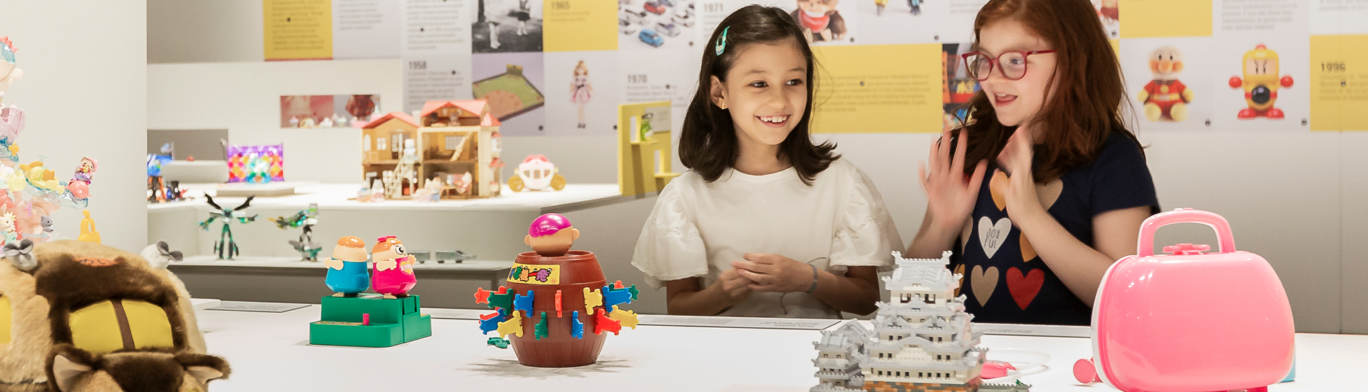 Duas meninas olhando os brinquedos expostos sobre mesa branca na exposição ‘Dōshin: os encantos dos brinquedos japoneses’, em cartaz na JHSP.
