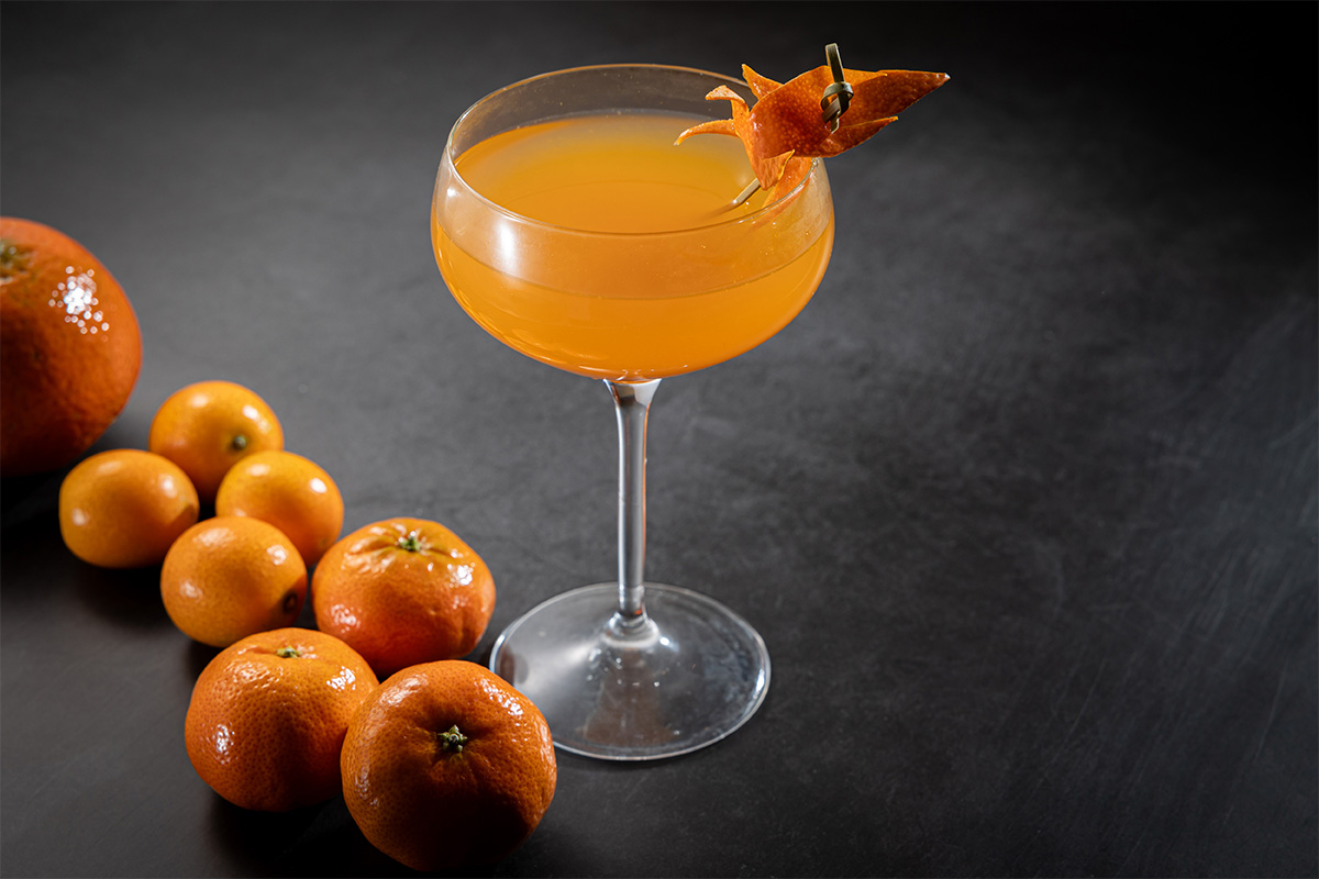 Fotografia de taça de drinque laranja com tangerinas dispostas na mesa.