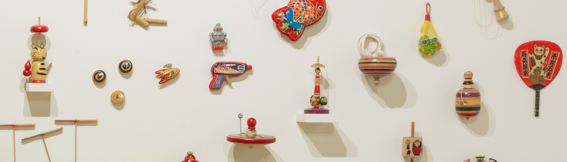 Brinquedos expostos em parede branca na exposição 'Doshin: os encantos dos brinquedos japoneses'