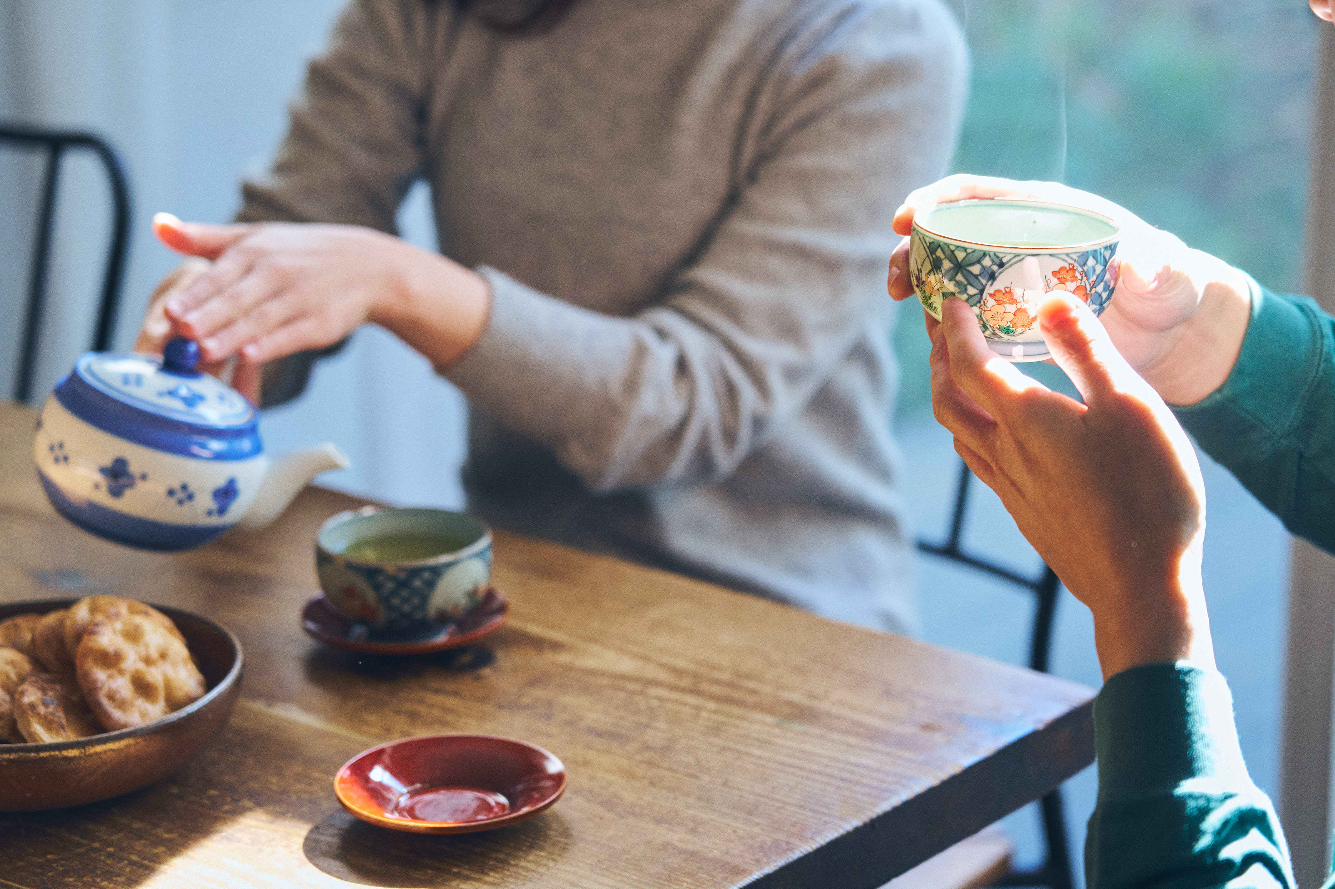 Foto com detalhes de mão segurando um copo de chá verde de cerâmica, e outra servindo o chá verde em cima de uma mesa.